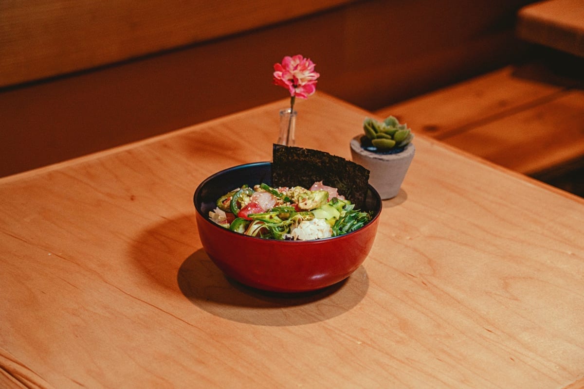 Poké bowl with greens and avocado in a red bowl, a pink flower behind it